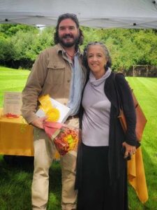 Will O'Brien dressed in khaki, holding a boquet of flowers standing next to Suzanne Gaetjens-Oleson, NHS CEO. Behind them is a table draped in a yellow tablecloth, and a pop-up tent. They are standing in a grass field.